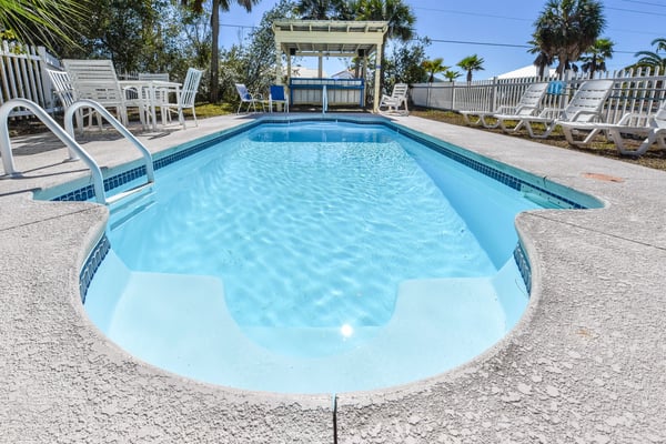 Rectangular swimming pool with white lounge chairs and pergola, surrounded by white fence and palm trees