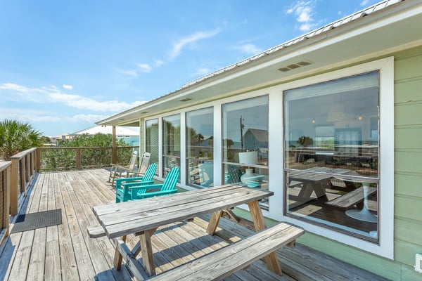 Elevated wooden deck with picnic tables and turquoise Adirondack chairs, overlooking coastal neighborhood with palm trees