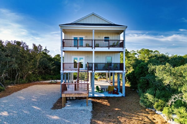 Three-story elevated beach house with white siding, multiple decks, and gravel driveway surrounded by trees