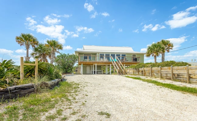 Elevated beach house with pale green exterior, wooden deck, and stairs, surrounded by palm trees and sandy driveway
